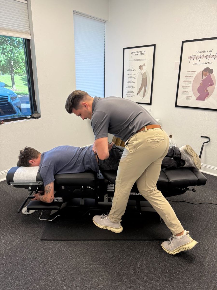 A chiropractor adjusts a patient's lower back as the patient lies face down on a treatment table in a clinic room.