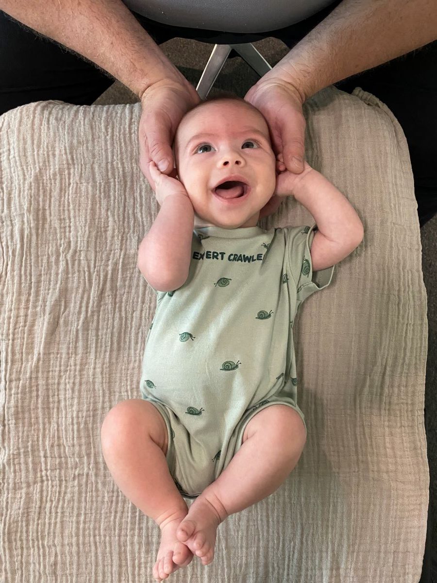 A smiling baby lies on a beige blanket, wearing a green onesie, with an adult's hands gently holding the baby's head—evoking a calming moment reminiscent of the gentle touch used in spinal decompression therapy.