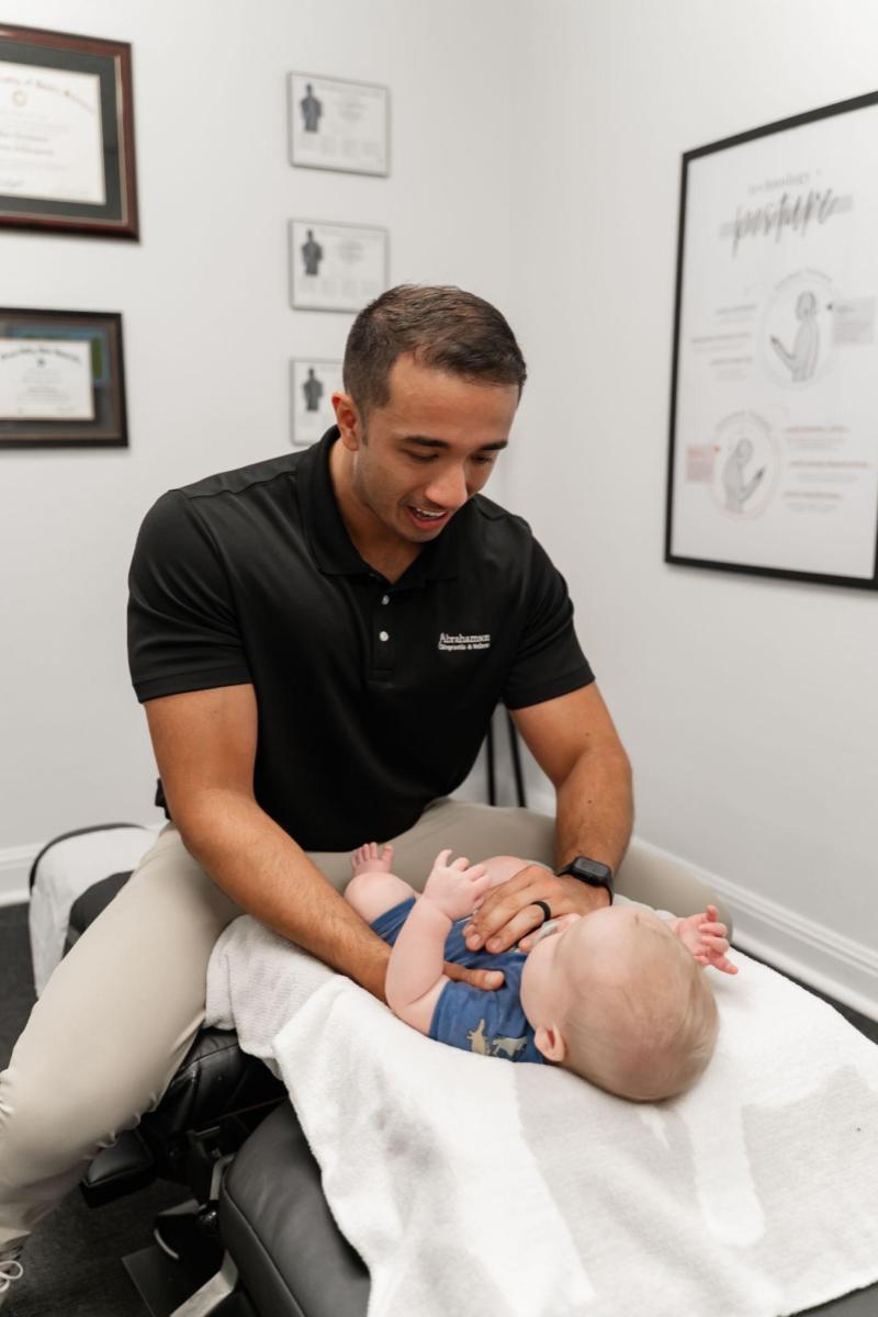 A man in a black polo shirt examines a baby lying on a towel in a medical office, with framed certificates and posters visible on the white walls.