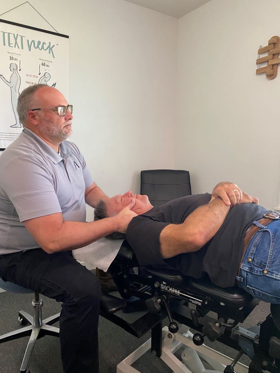 A chiropractor near me accepting new patients adjusts a patient's neck as the patient lies on an exam table in a clinic room. A poster about "text neck" is visible on the wall in the background.