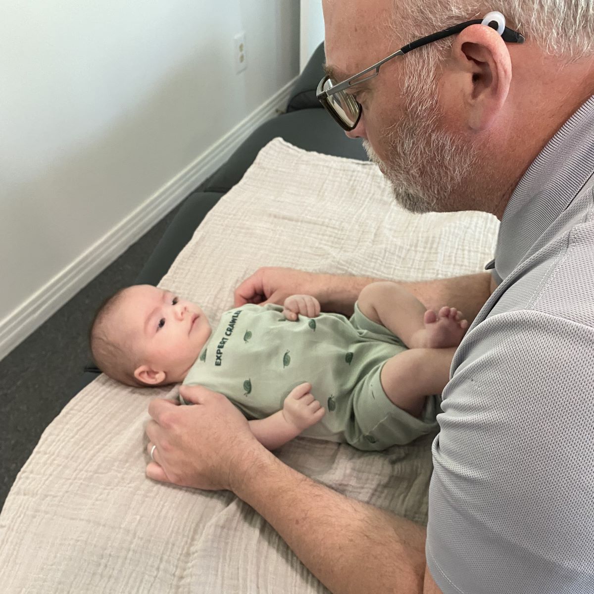 An adult with glasses gently holds a baby lying on a blanket, as they look at each other in a well-lit room—creating a calm scene reminiscent of the care and connection often found at a Sumner chiropractor’s office.